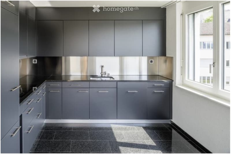 Modern kitchen with grey cabinets, stainless steel backsplash, and a black countertop. Features a sink with a modern faucet and ample cabinet space.
