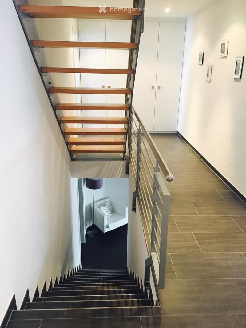 Modern staircase with wooden steps and metal railing, leading to a lower level with a white armchair and floor lamp visible below.