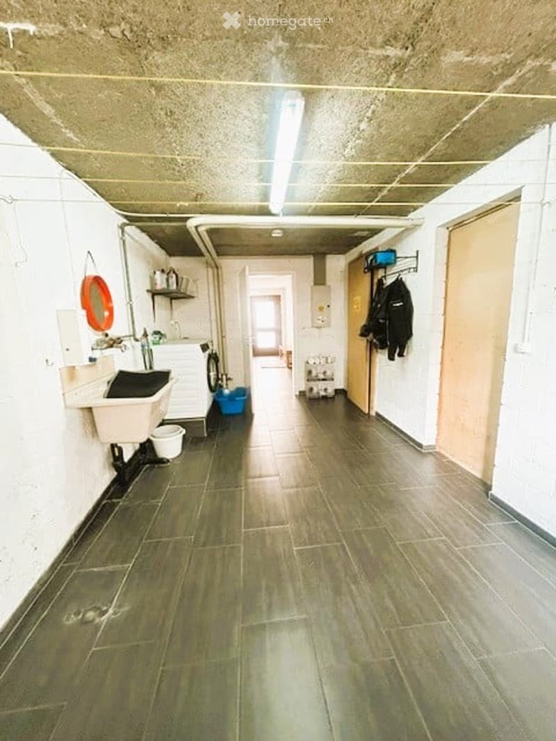 A narrow hallway with tiled flooring, white walls, and a concrete ceiling. There are various items visible, including a sink, shelves, a washing machine, and a coat rack with jackets.