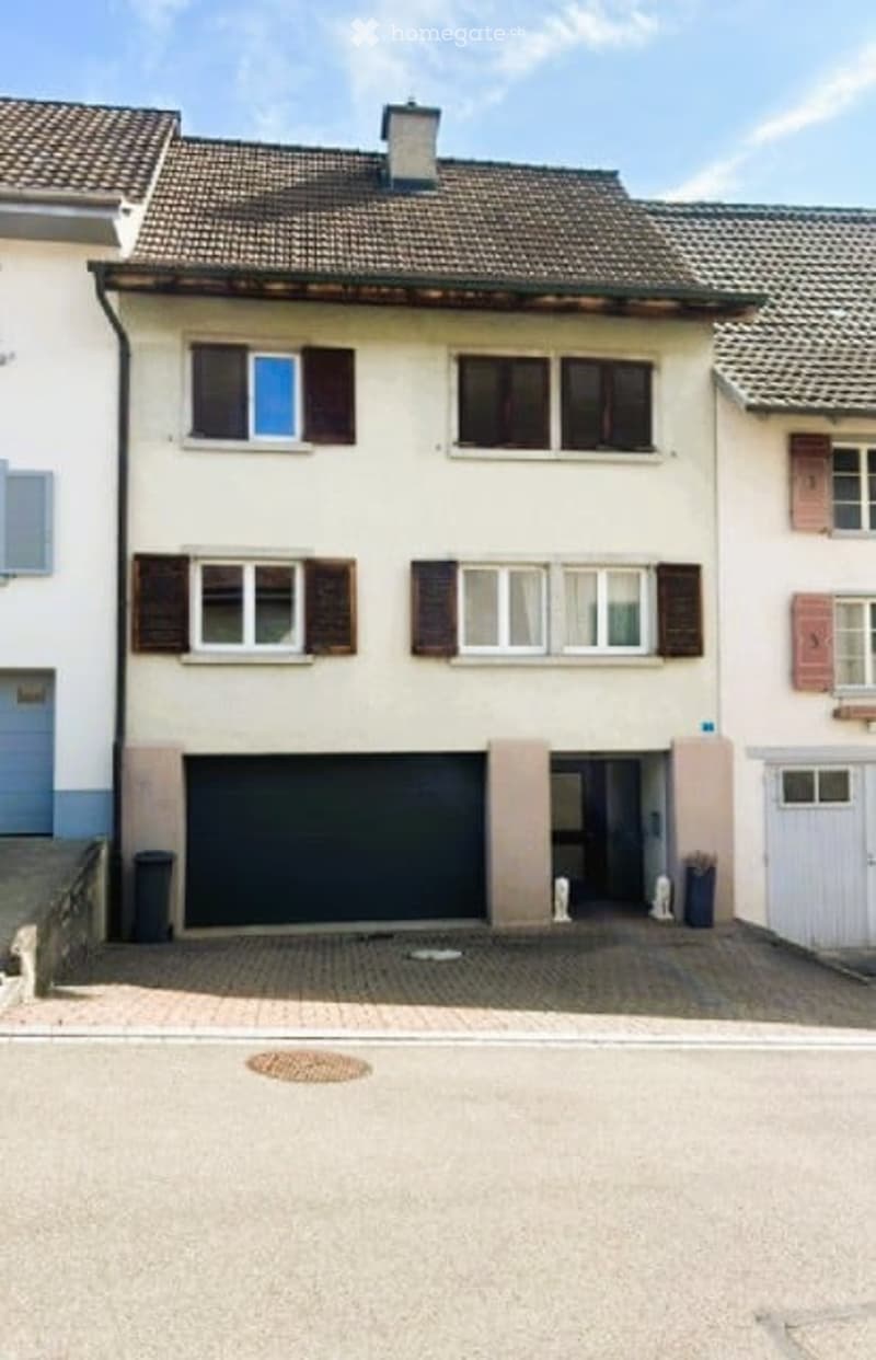 Two-story house with beige facade, multiple windows with shutters, and a garage entrance.
