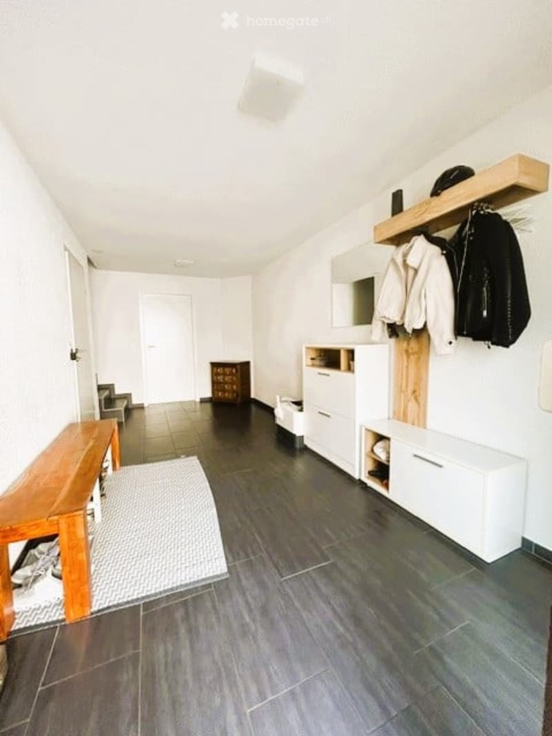 A modern hallway with a wooden bench, a coat rack with jackets, a white storage unit, and a small cabinet. The floor is dark wood, and the walls are white.