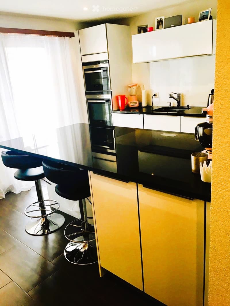 Modern kitchen with white and yellow cabinets, black countertops, stainless steel appliances, and two black bar stools.