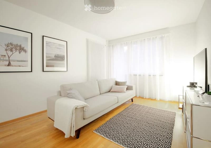 Modern living room with white walls, wooden floor, large windows with white curtains, a white sofa with pillows, a coffee table, a mirror, and two picture frames on the wall.