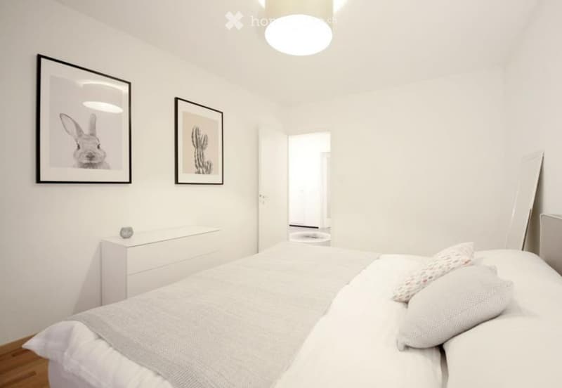 Modern, minimalist bedroom with a white bed, nightstand, and framed artwork on the walls.