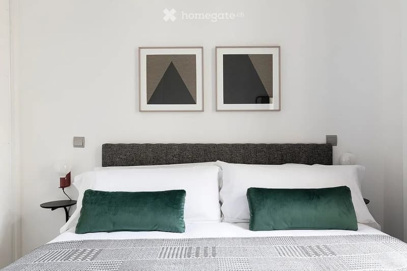 Modern bedroom with a neatly made bed, white pillows, green throw pillows, a grey headboard, and two framed wall artworks above the bed.