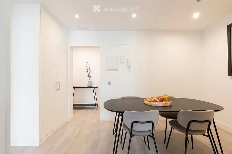 Modern dining area with a round wooden dining table, four chairs, a small side table with a vase, and a tray with food and drinks.