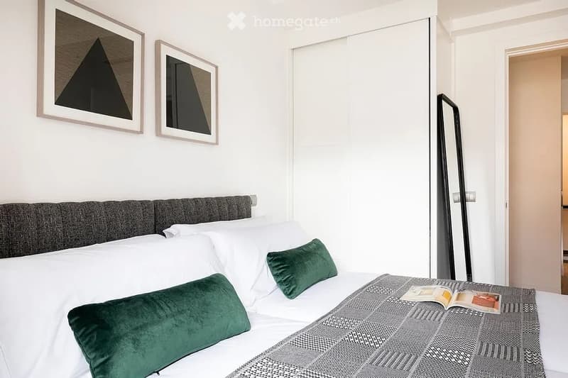 Modern bedroom with a large bed, grey headboard, white pillows, green throw pillows, grey and white patterned blanket, and two framed artworks on the wall.