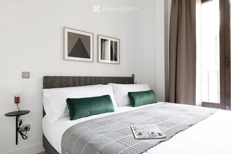 Modern bedroom with a neatly made double bed, green accent pillows, a grey headboard, wall-mounted nightstand with a lamp, and decorative mirrors on the wall.