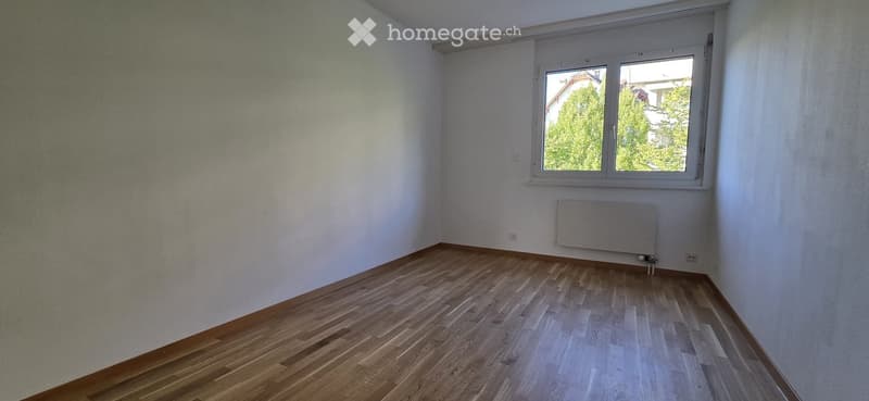 Empty room with wooden flooring, white walls, and a window with a view of greenery outside. A radiator is mounted on the wall below the window.
