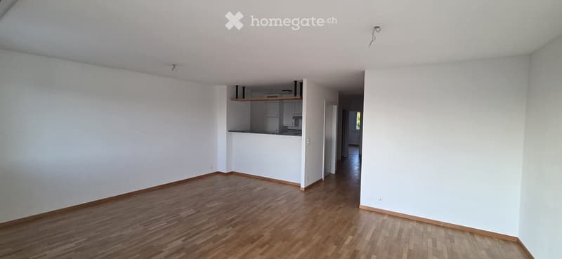 Empty room with wooden flooring and white walls, featuring an open kitchen area with cabinets and appliances visible in the background.