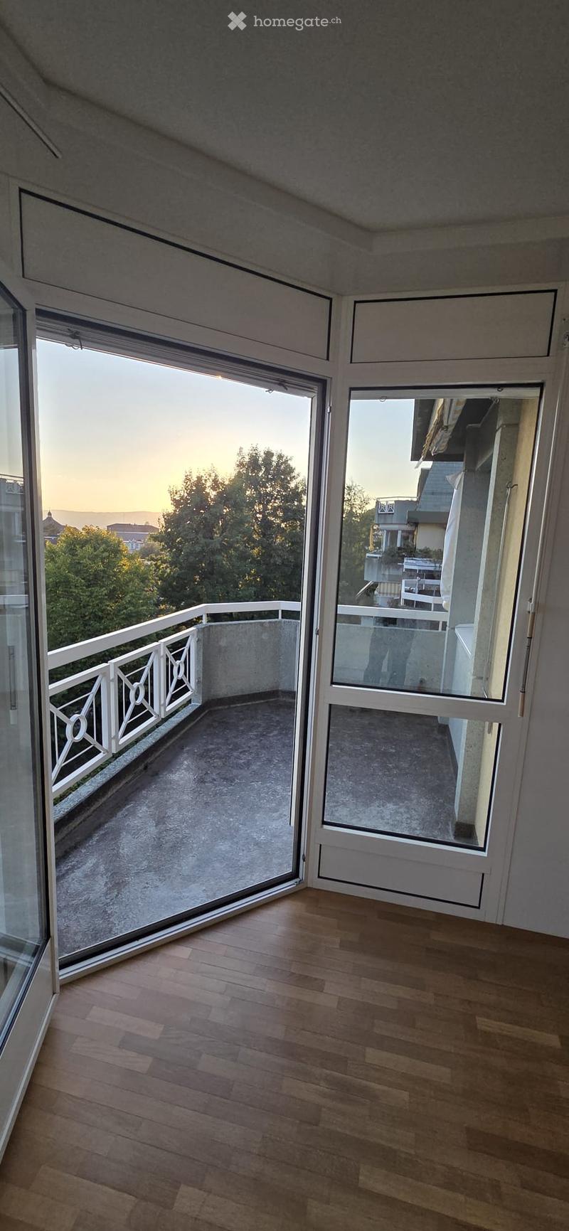 Empty room with wooden flooring, large sliding glass doors leading to a balcony with white railings and a view of trees and buildings.
