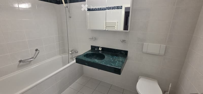 Modern bathroom with white tiled walls and floor, featuring a bathtub, a green marble sink, a toilet, a mirror, and a handrail for accessibility.