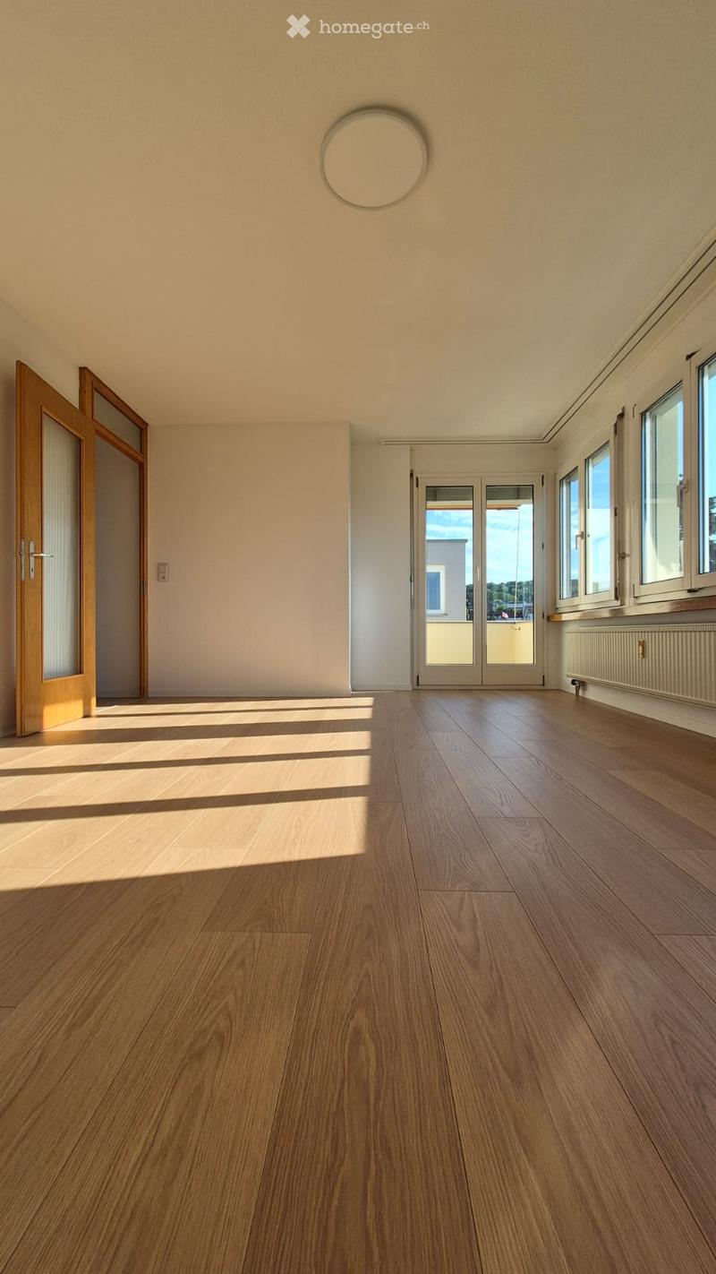 Empty room with wooden flooring, large windows, and a sliding glass door leading to a balcony.