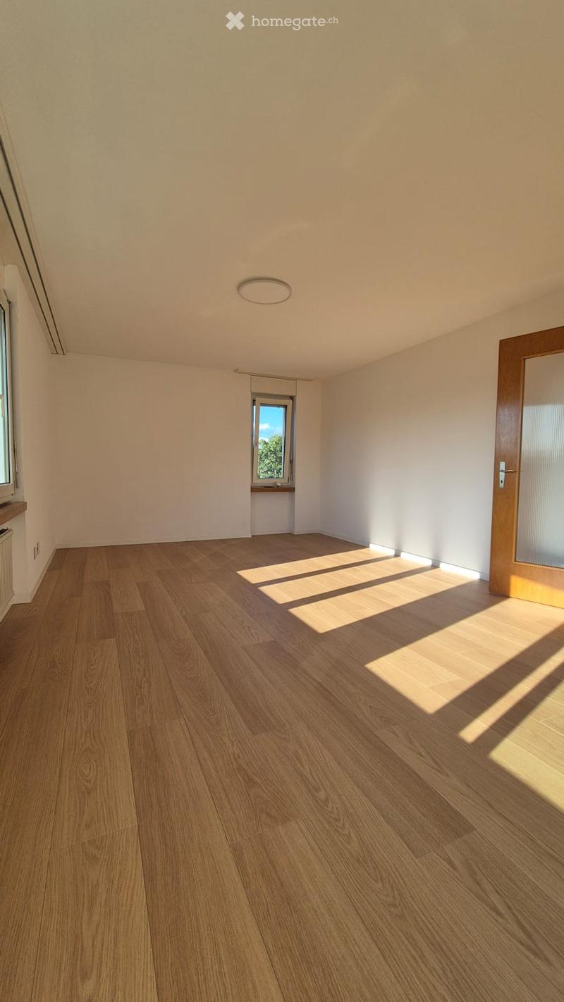 Empty room with light wooden flooring, white walls, and large windows allowing natural light to enter. The room features a simple, modern design with a recessed ceiling light and a door leading to another space.