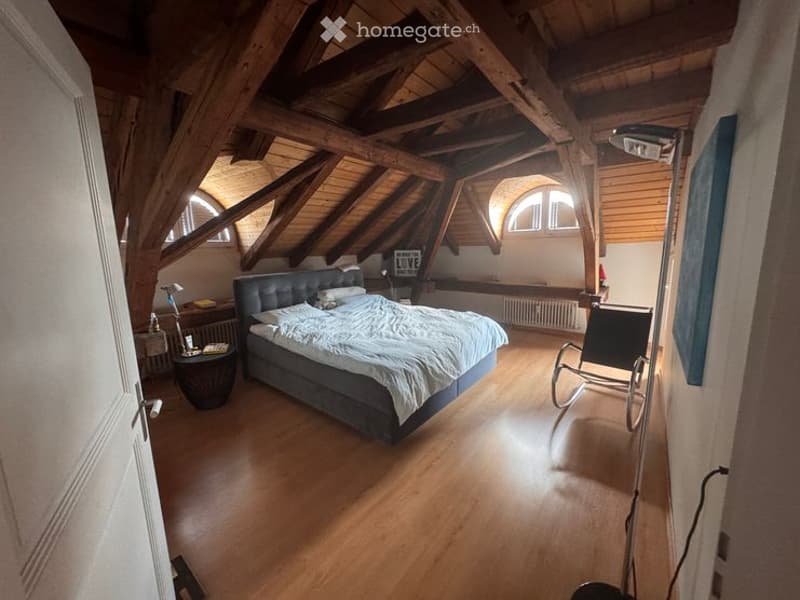 Cozy attic bedroom with wooden beams, exposed wooden ceiling, large windows, a dark blue bed, nightstand, and a wicker chair.