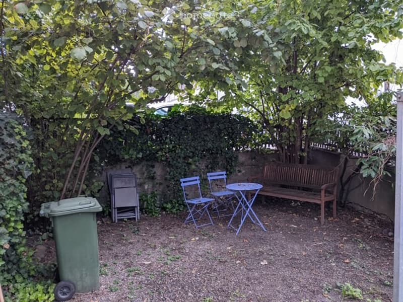 Small, enclosed garden with a wooden bench, two blue folding chairs, a round table, a green trash bin, and a metal folding chair. The area is surrounded by a hedge and trees, creating a private outdoor space.