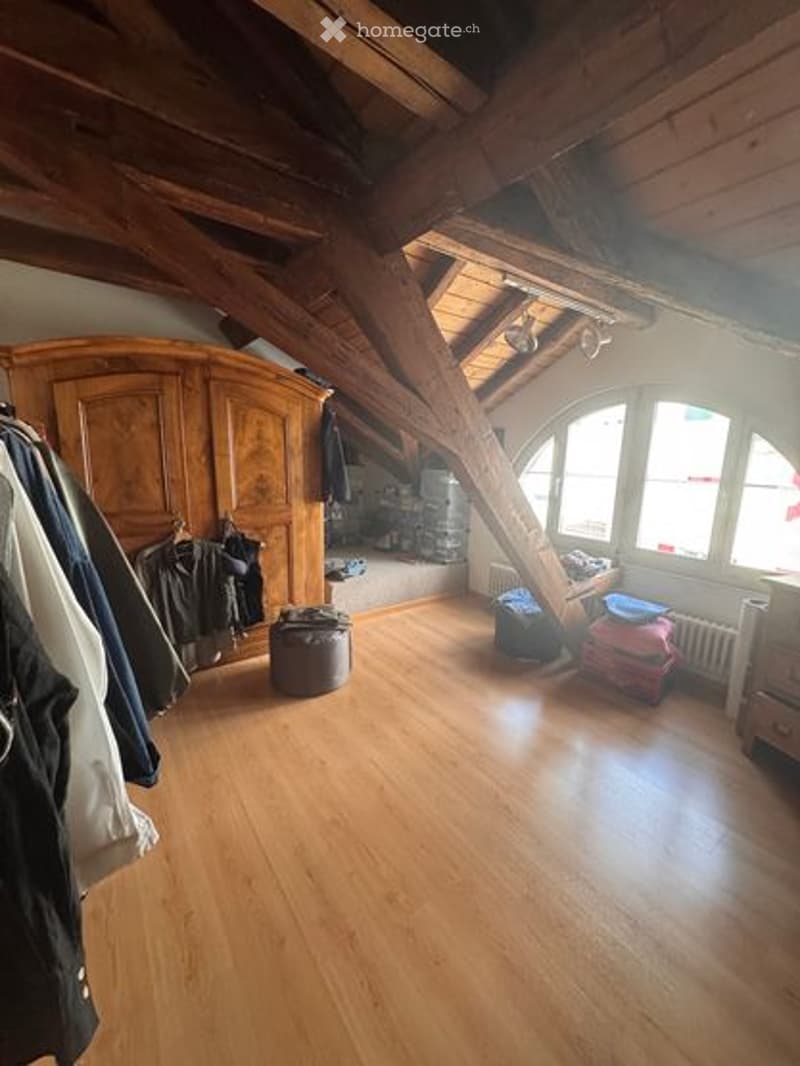Loft-style room with wooden beams, exposed wooden ceiling, large arched windows, hardwood floor, wardrobe, stool, radiator, and drawer unit.