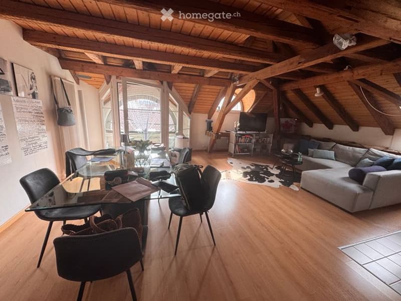 Bright living room with wooden beams, hardwood flooring, large windows, a grey sectional sofa, coffee table, glass dining table with chairs, TV, bookshelf, and a cowhide rug.