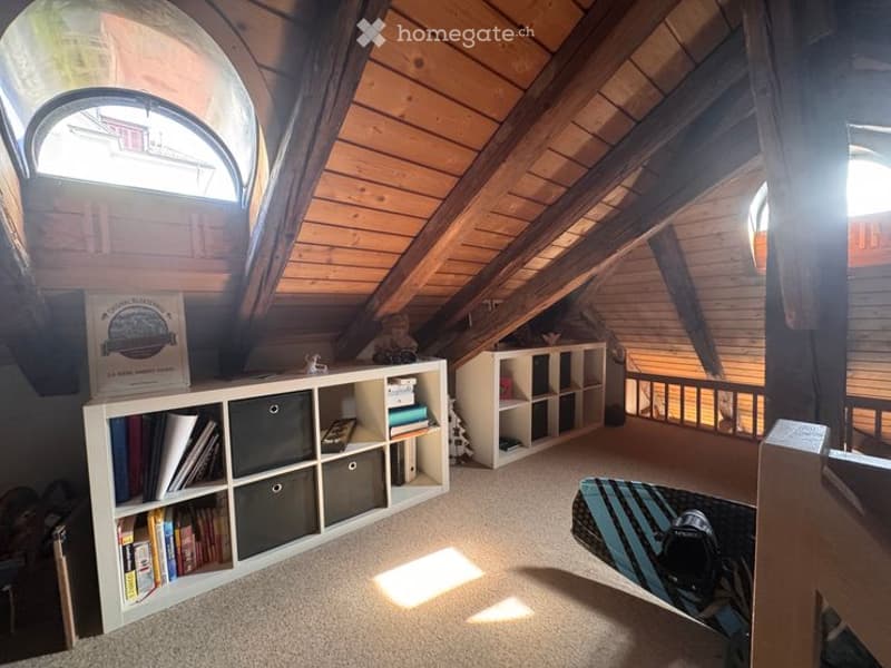 Cozy attic room with wooden slanted ceilings, large skylight windows, built-in white shelving units filled with books and storage bins, a small toy on the shelf, and a striped rug on the floor.
