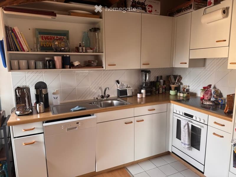 A modern kitchen with white cabinets, wooden countertops, and various appliances including a sink, dishwasher, oven, coffee maker, and blender. Shelves hold books, cups, and decorative items.