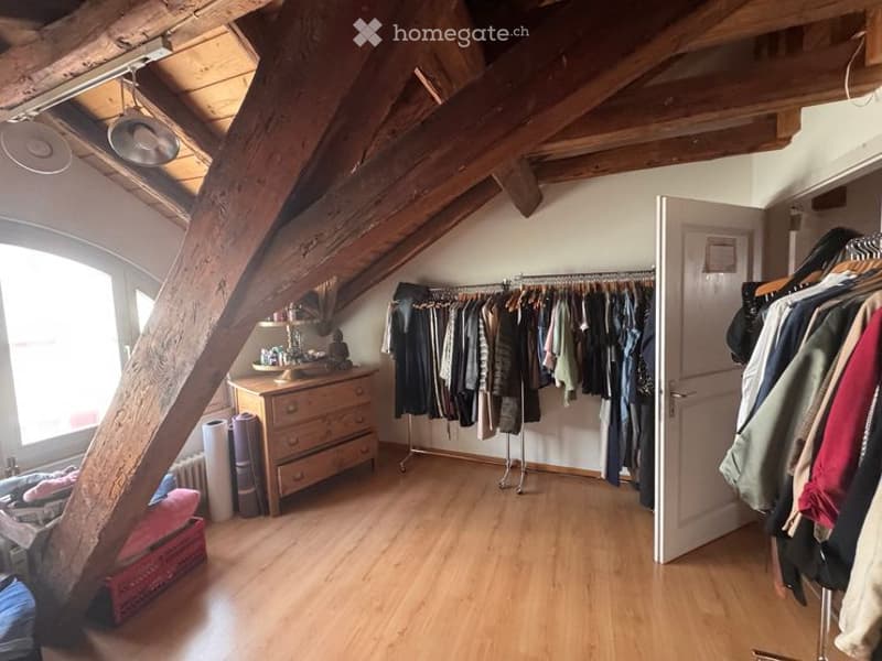 Attic room with wooden beams and exposed ceiling, featuring a wooden floor, a dresser, a clothing rack, and a wardrobe. The room has large windows allowing natural light to fill the space.
