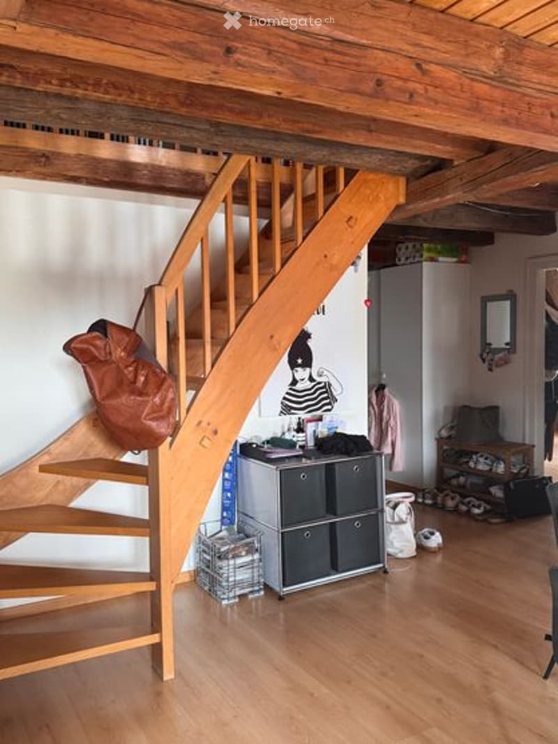 A cozy, open-plan living space with exposed wooden beams and a wooden staircase leading to an upper level. The room features a modern storage unit, a refrigerator, a mirror, a chair, a basket, a stool, and a painting on the wall.