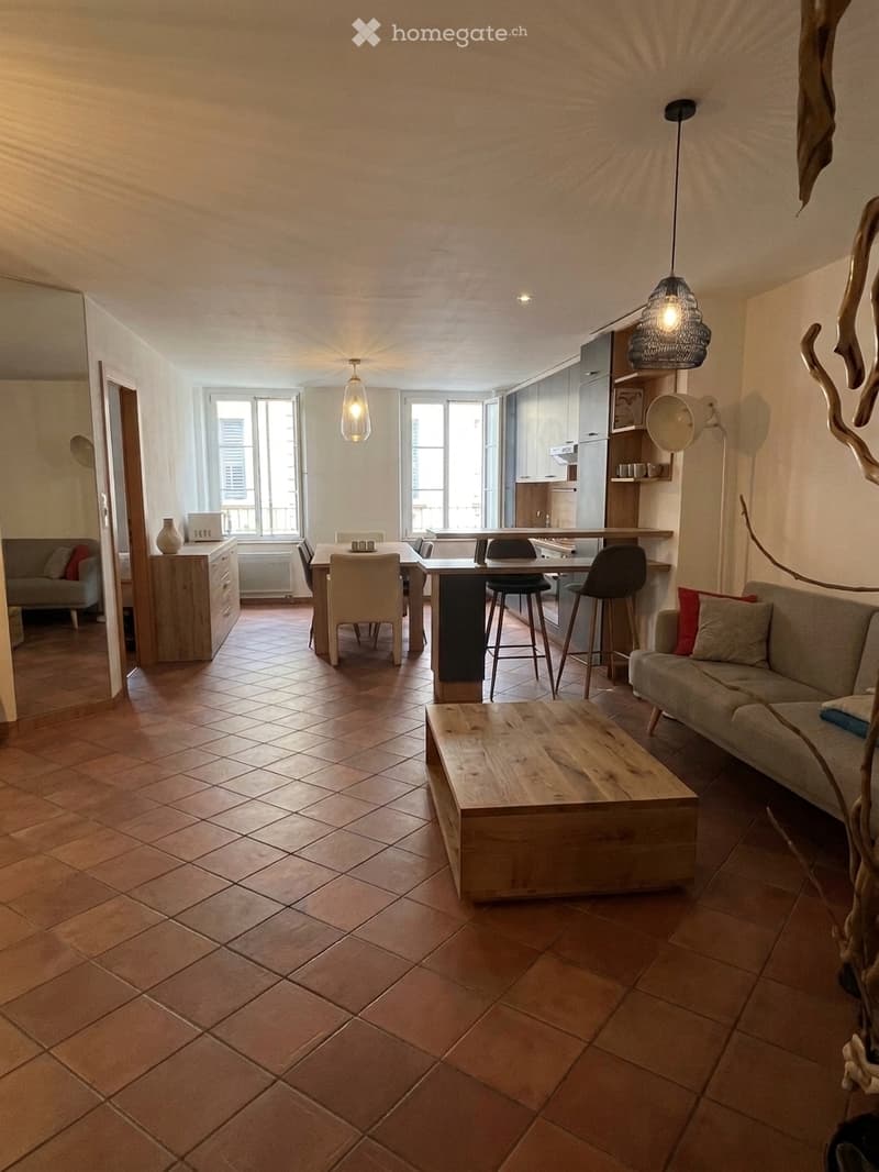 Open-plan living and dining area with tiled flooring, large windows, a grey sofa, wooden coffee table, dining table with chairs, and kitchen area with cabinets.