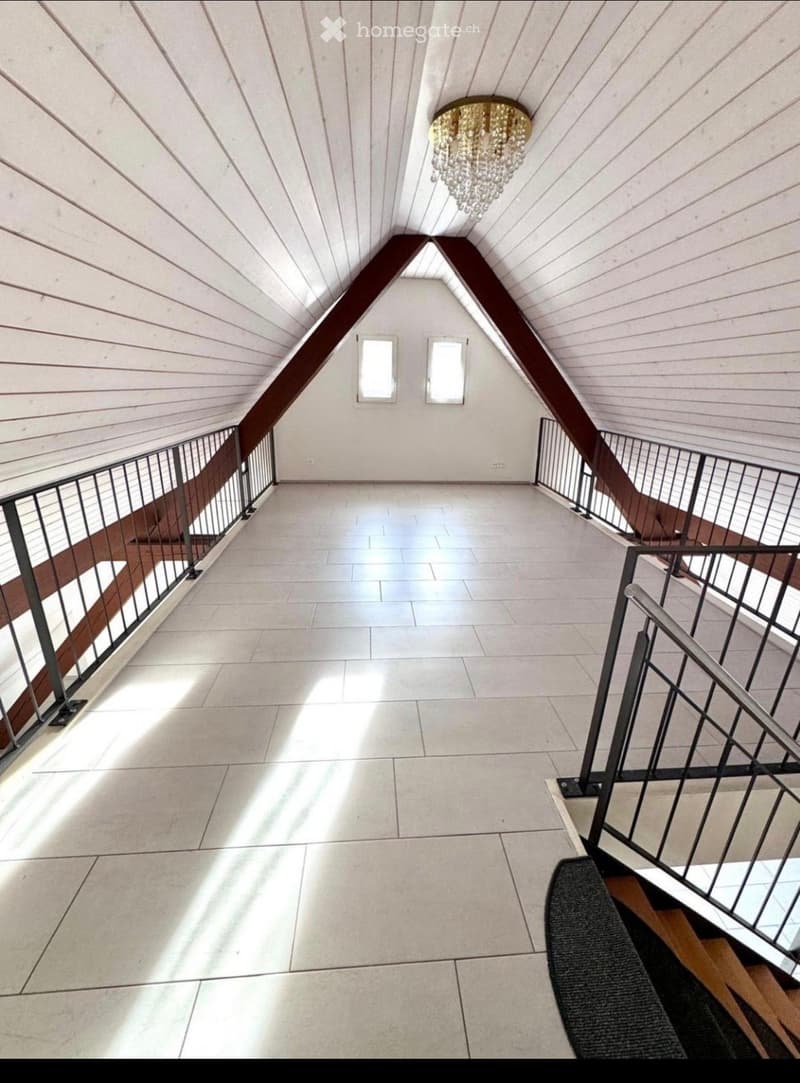 Bright, open hallway with a high, slanted wooden ceiling, large windows, and a chandelier. The floor is tiled, and there are metal railings on both sides leading to stairs.