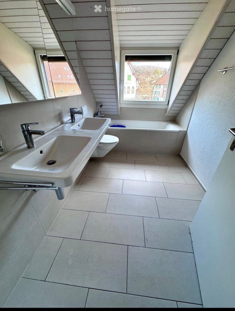 Modern bathroom with two sinks, a toilet, and a bathtub, featuring light-colored tiles and wooden paneling on the walls.
