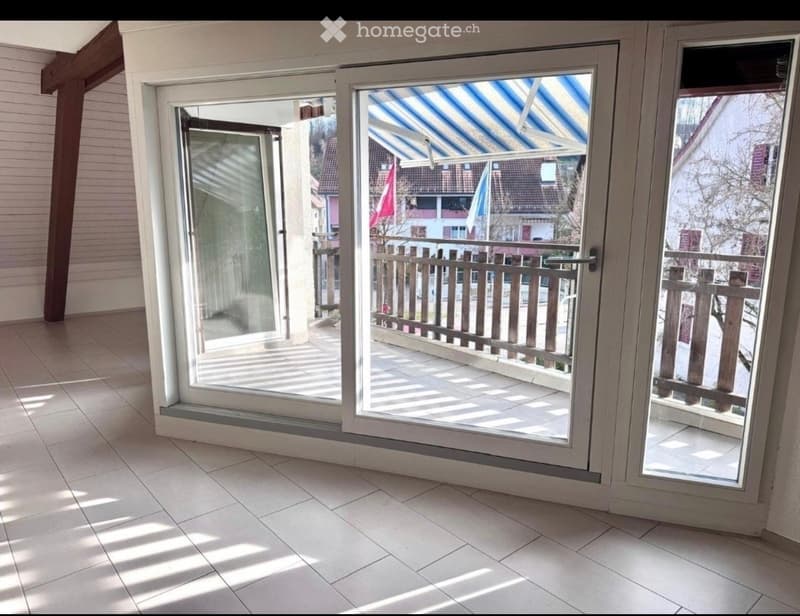 View of a tiled indoor space with large sliding glass doors leading to a covered outdoor balcony area with wooden railings.