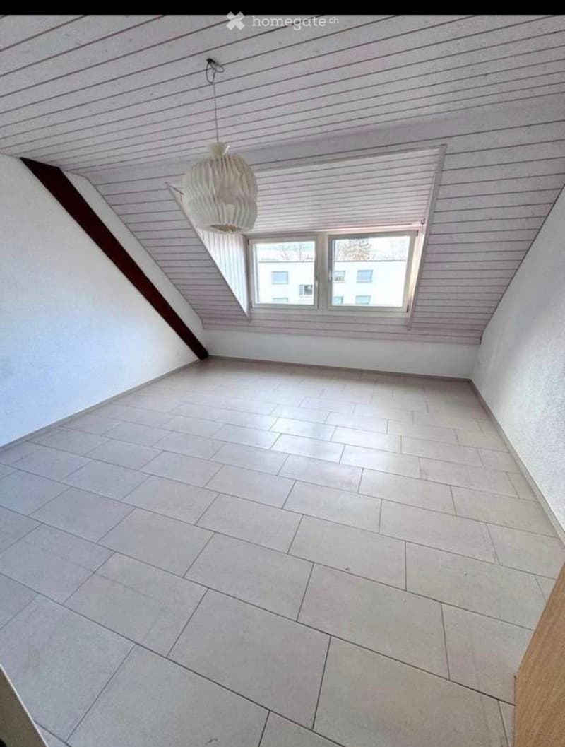 Empty attic space with wooden slatted ceiling, white tiled floor, and a single window with white frame showing a neighboring building.