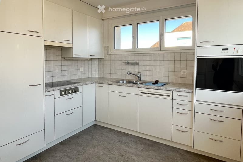 Modern kitchen with white cabinets, granite countertops, stainless steel sink, oven, and dishwasher. Three windows provide natural light.