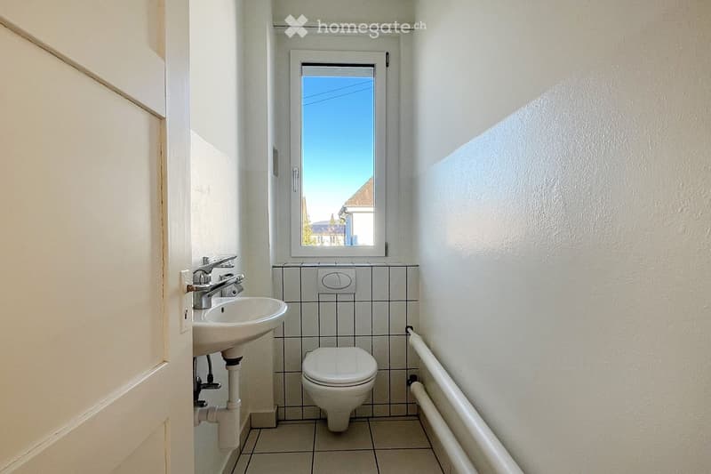Small bathroom with a white tiled wall, a sink with a chrome faucet, and a toilet. A window with a view of the outside is above the toilet.