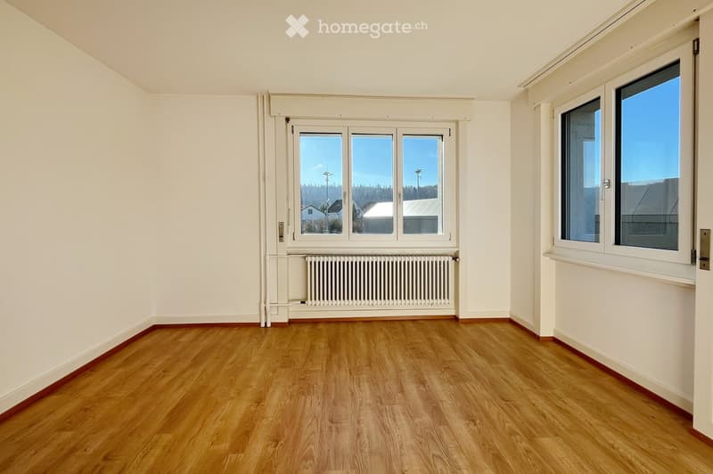 Empty room with wooden flooring, white walls, and large windows offering a view of the outside. A radiator is visible under one of the windows.
