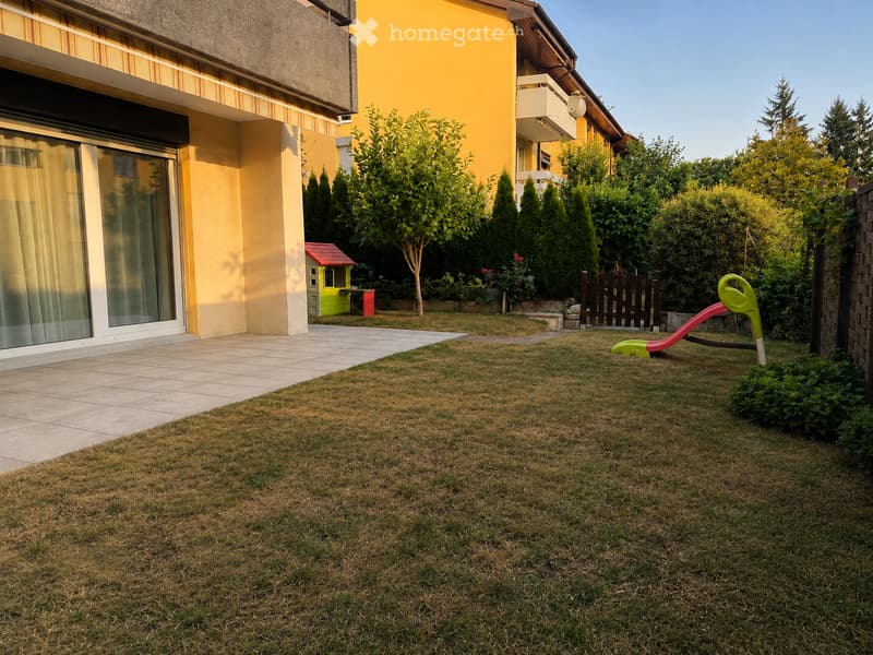 Well-maintained garden with green grass, a small slide, a playhouse, and surrounding trees and shrubs. The house features large windows and a balcony.