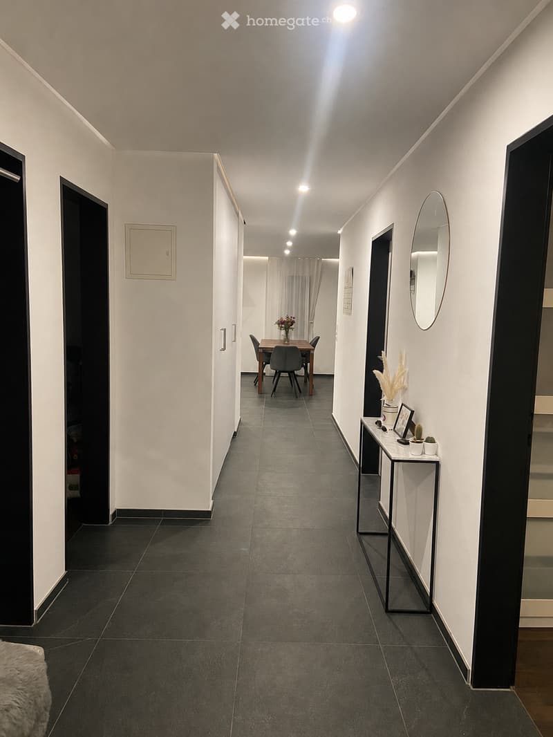 Modern hallway with dark tiled flooring, white walls, and recessed lighting. Features a small dining area with a table and chairs, a side table with decorative items, and a mirror on the wall.