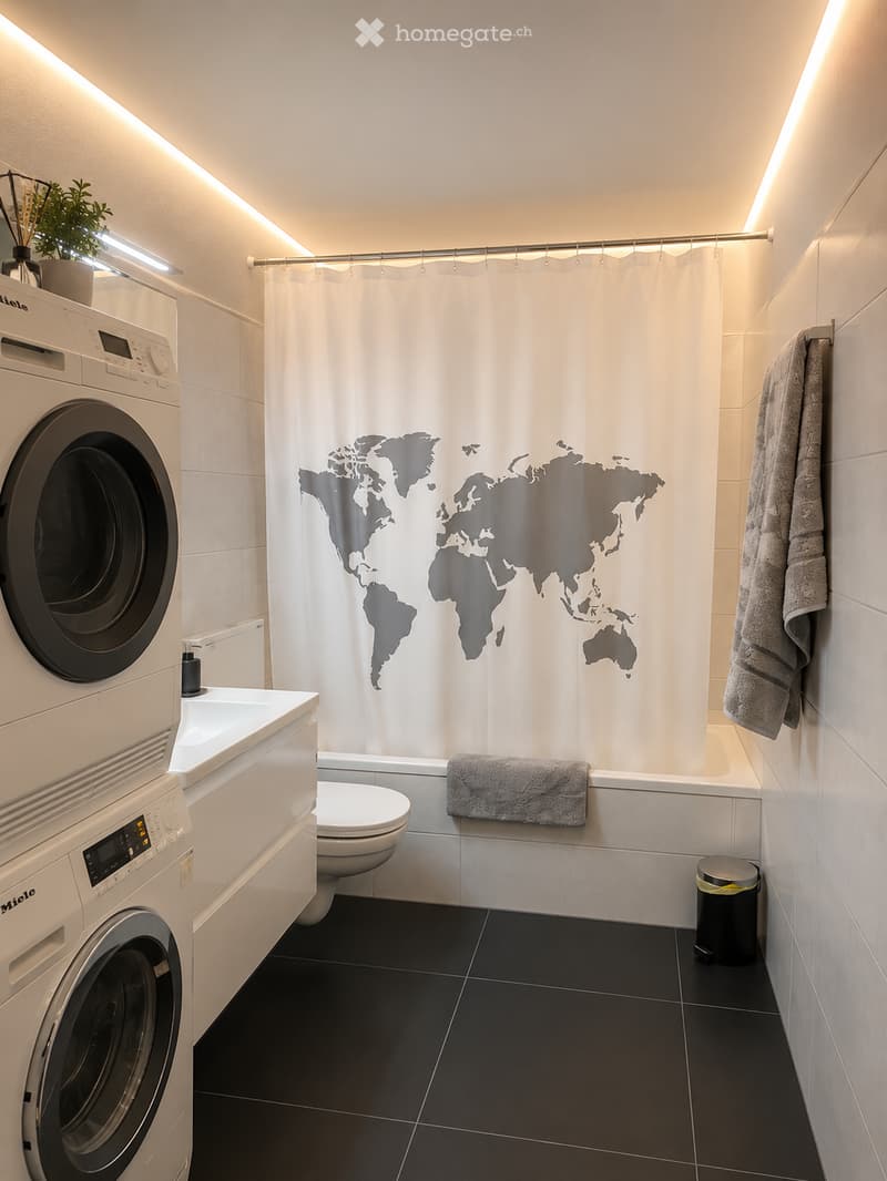 Modern bathroom with black tiled floor, white walls, washing machine, toilet, bathtub, shower curtain with world map design, towels, and a small plant on the washing machine.