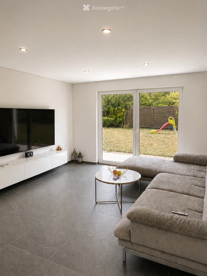 Modern living room with a grey sectional sofa, glass coffee table, flat-screen TV on a white cabinet, and a view of a garden through large sliding doors.