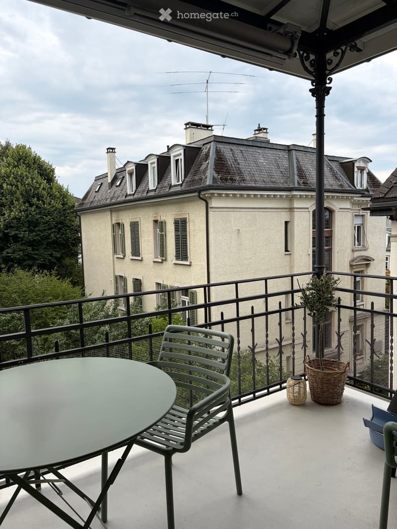Balcony with a round table, two chairs, and potted plants, overlooking a classic European building with a mansard roof.