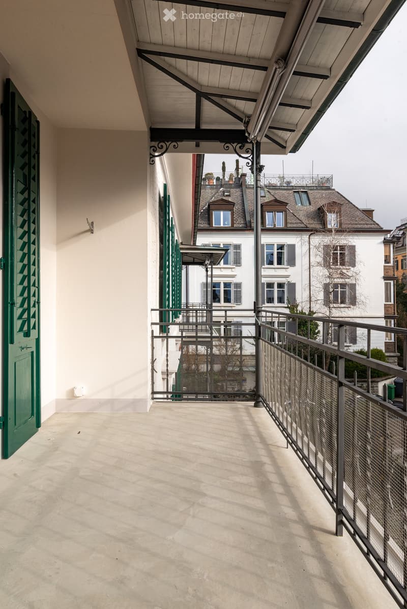 Balcony with a metal railing, concrete floor, and a green door leading to the interior. The view shows neighboring buildings with classic architecture.