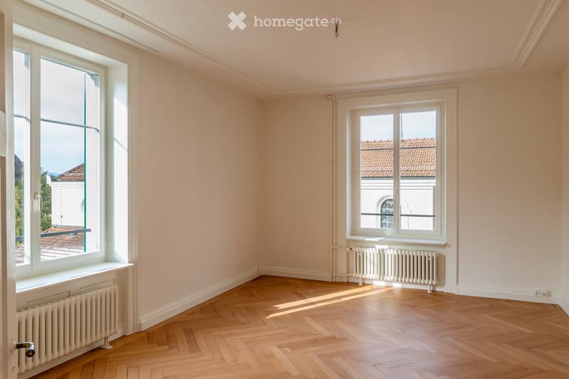 Empty room with light-colored walls, wooden herringbone flooring, and large windows offering views of rooftops and buildings.