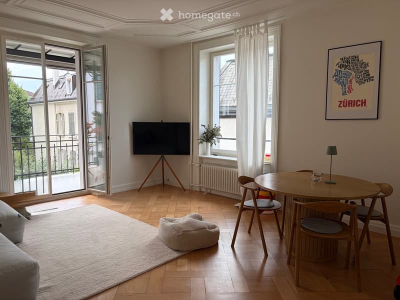Bright living room with wooden flooring, large windows, balcony access, sofa, coffee table, dining table with chairs, lamp, television, and a potted plant on the windowsill.