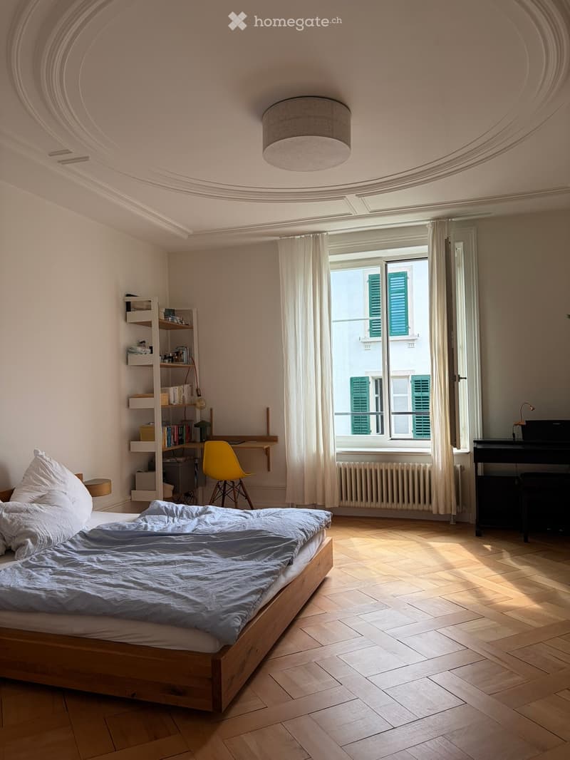 Bright bedroom with wooden flooring, large window with white curtains, bed with light blue bedding, yellow chair, shelf with books, desk, and lamp.