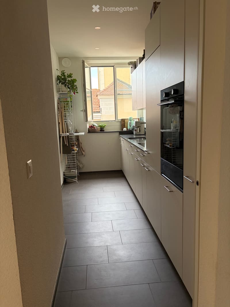 Modern kitchen with grey tiled floor, white cabinets, built-in oven, sink, and small plant on the windowsill.