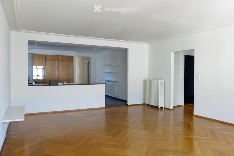 An empty room with wooden flooring, white walls, and a white radiator. A kitchen area is visible in the background with wooden cabinets and a stainless steel oven.