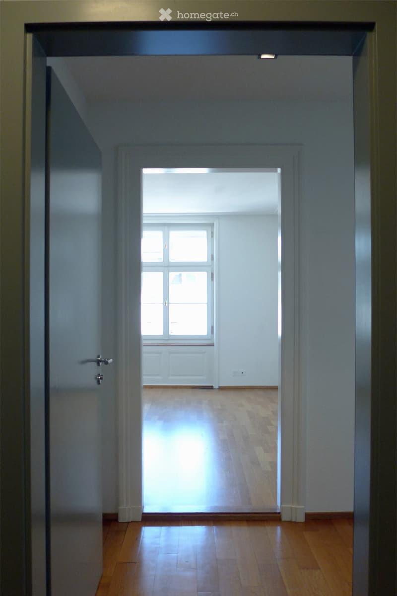 Empty hallway with wooden flooring, white walls, and a door leading to a brightly lit room with large windows.