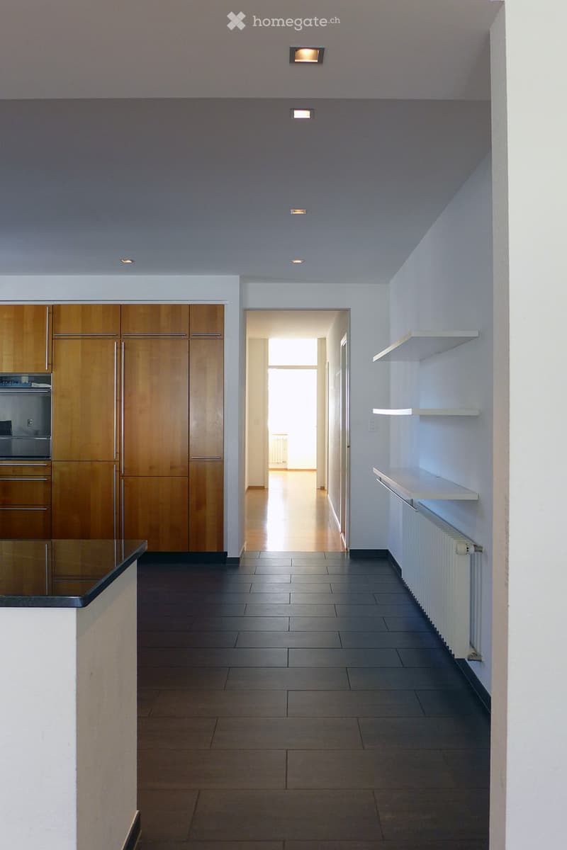 Empty hallway with tiled floor, wooden cabinets on one side, white shelves on the other, and a doorway leading to another room.
