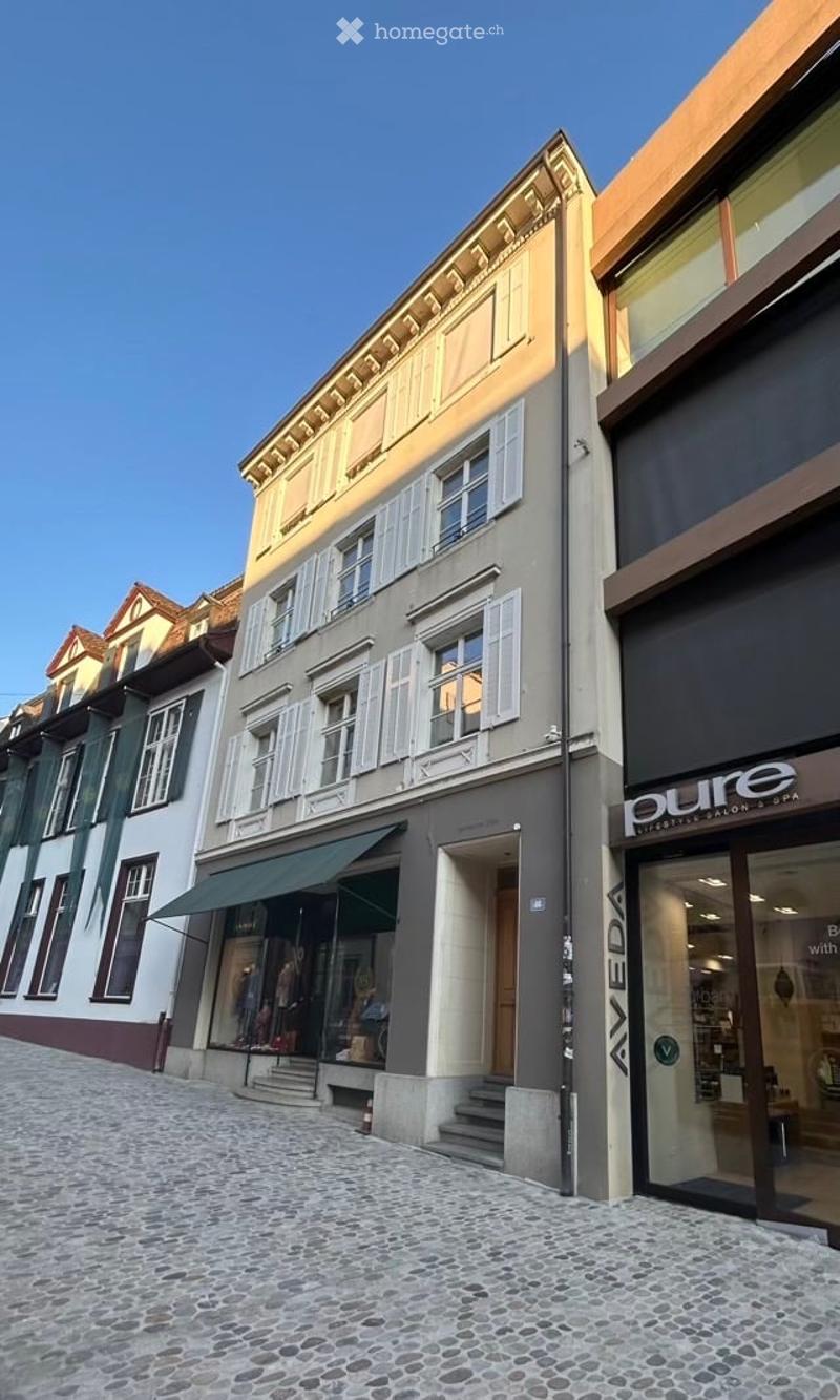 A multi-story building with classic European architecture, featuring a mix of beige and grey facades, white shutters, and a green awning over the entrance. The building is part of a row of similar structures on a cobblestone street.