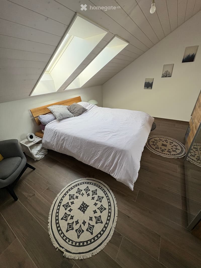 Cozy attic bedroom with wooden slanted ceilings, skylights, a large bed with pillows and blankets, a nightstand with a lamp, a round patterned rug, and a grey armchair.