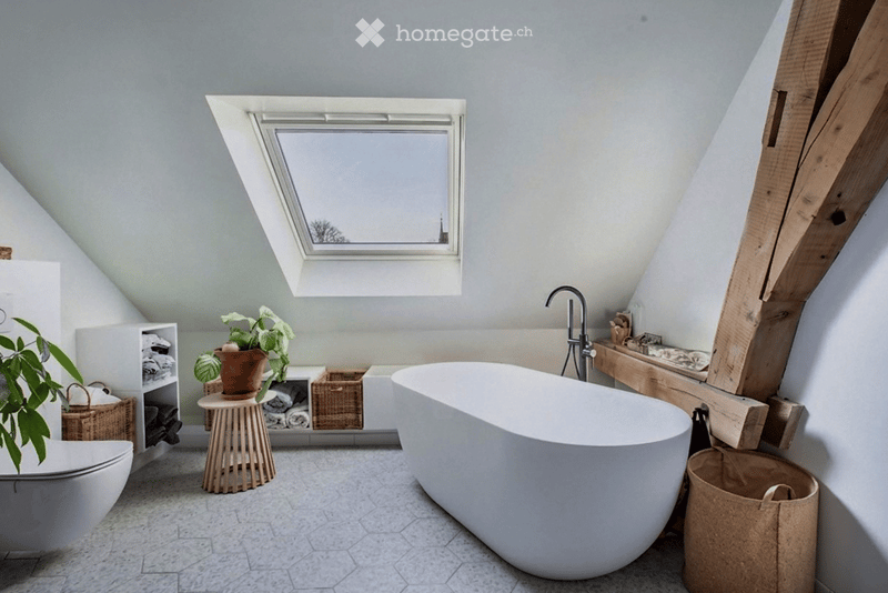 Modern bathroom with a freestanding bathtub, toilet, small wooden stool with a potted plant, and a shelf with towels and baskets. The room features a skylight window and exposed wooden beams.